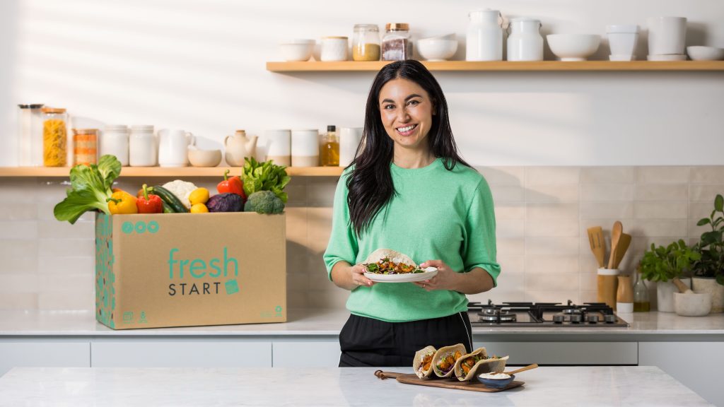 Nadia Lim holding healthy tacos standing next to a Fresh Start box packed with fresh vegetables and produce on a kitchen bench